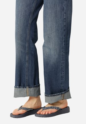 Des pieds portant des tongs tissées bleu marine avec des brides florales, associés à un jean en denim bleu retroussé contre un fond blanc uni.