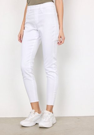 Kobieta ubrana w dopasowane białe spodnie o długości do kostek oraz białe sneakersy, stojąca na tle gładkiego białego tła.