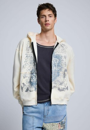 Joven con una sudadera con capucha crema con cremallera, con letras y patrones azules, una camiseta negra y vaqueros claros de retales.