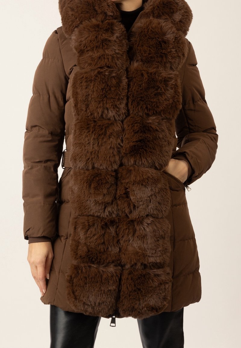 Femme portant un manteau matelassé marron avec une large bordure en fourrure douce et foncée le long de l'avant et autour du col, les mains dans les poches.
