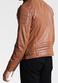 Giacca in pelle marrone con dettagli trapuntati sulle spalle, forma aderente, chiusura con zip e accentuazione con fibbia laterale. Texture liscia con una leggera lucentezza.