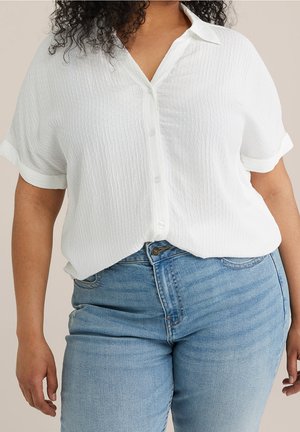 Kvinde iført tekstureret hvid kortærmet skjorte med knapper, stoppet ind i lyseblå højtaljede jeans, stående mod en neutral baggrund.