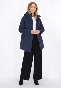 Jachetă bleumarin, cu fermoar și glugă. Fabricată dintr-un material neted, are detalii cu nasturi tip clichet și un croi lejer, asortată cu pantaloni largi negri.