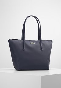 Bolso tipo tote de color azul marino, fabricado en un material texturizado, con dos asas y un cierre de cremallera, que presenta un pequeño logo verde de Lacoste en la parte frontal.