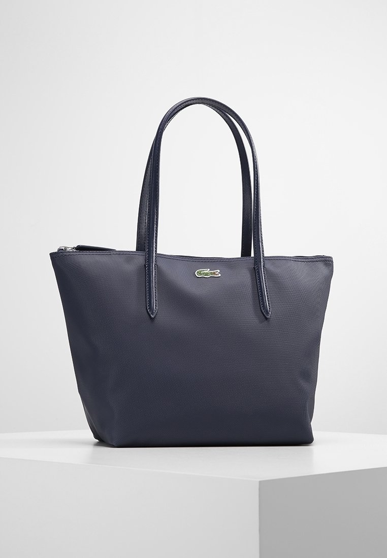 Bolso tipo tote de color azul marino, fabricado en un material texturizado, con dos asas y un cierre de cremallera, que presenta un pequeño logo verde de Lacoste en la parte frontal.