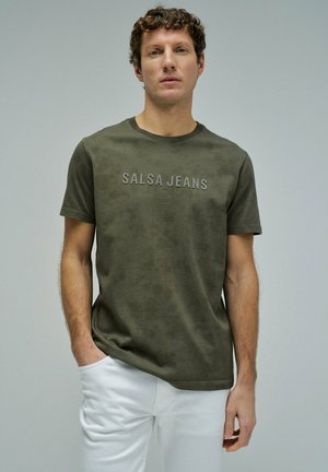 Hombre con cabello rizado viste una camiseta verde oliva con la inscripción "SALSA JEANS" y pantalones blancos, de pie contra un fondo gris liso.