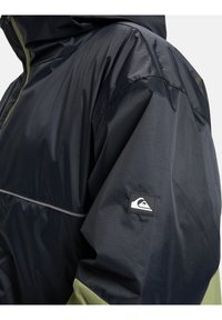 Navy and green water-resistant jacket with a zip, textured fabric, and logo patch on the sleeve; features a hood and reflective detailing.