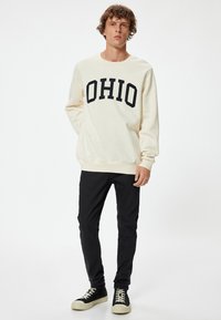 Sudadera crema con estampado negro "OHIO", puños y dobladillo de canalé. Combinada con pantalones negros ajustados y zapatillas negras con cordones blancos.
