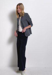 Frau mit blondem Haar, die einen grauen Blazer, ein weißes Top, schwarze, weite Hosen und weiße Sneakers trägt und an einer weißen Wand steht.