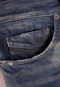Jeans i denim med en avbleknad blå tvätt, slitna detaljer och en framficka som visar en svart etikett med varumärket.