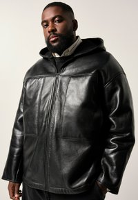 Uomo con barba che indossa un hoodie in pelle nera con zip sopra una camicia con colletto e un maglione, in piedi con una mano in tasca contro uno sfondo neutro.