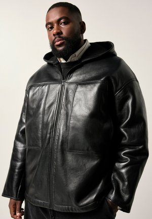 Man with beard wearing a black leather zip-up hoodie over a collared shirt and sweater, standing with one hand in pocket against a neutral background.