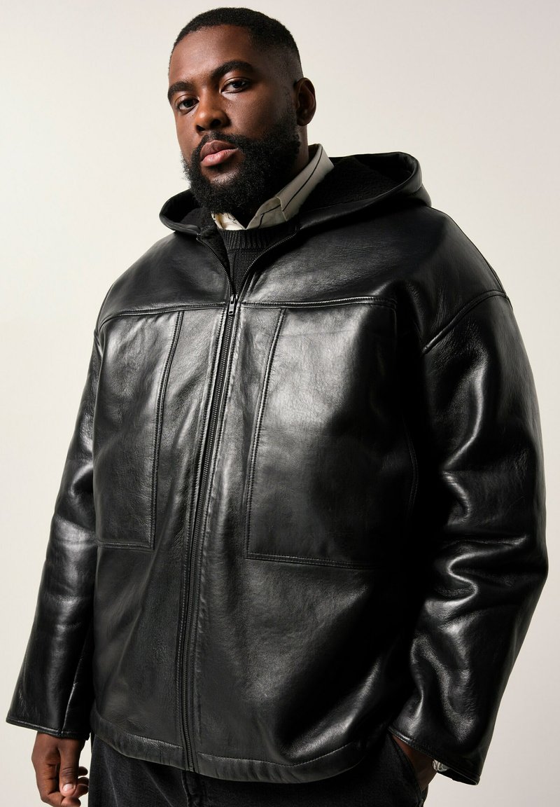 Man met een baard, gekleed in een zwart leren hoodie met rits over een overhemd en trui, staand met één hand in zijn broekzak tegen een neutrale achtergrond.