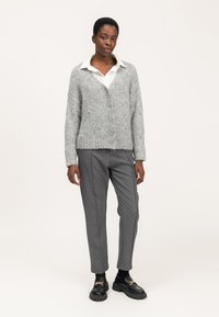 Cárdigan gris de punto con botones, llevado sobre una camisa blanca, combinado con pantalones gris a rayas con puntos blancos. Mocasines negros gruesos.