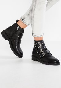 Černé kožené kotníkové boty se stříbrnými sponami a nýty. Mají boční zip a texturovanou podrážku pro lepší trakci.