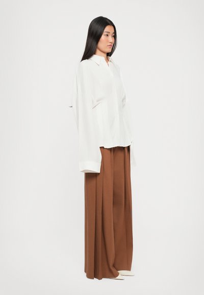 White oversized shirt with a collared neckline, paired with wide brown trousers. Smooth fabric and relaxed fit enhance overall silhouette.