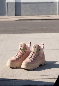 Paire de bottes épaisses rose clair avec semelles épaisses et lacets verts posées sur un trottoir en béton ensoleillé près d'une rue.