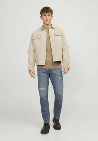 Chaqueta de mezclilla color beige claro con dos bolsillos frontales sobre una camisa marrón claro, combinada con jeans azules desgastados y botas oscuras de cordones.
