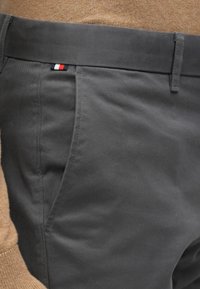 Pelēkas kokvilnas bikses ar gludu tekstūru, ar nepamanāmu sānu kabatu un mazmāju trīs krāsu logotipa etiķeti netālu no jostasvietas.