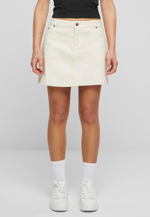 Mujer con una mini falda de mezclilla blanca, calcetines blancos, zapatillas blancas y una parte superior negra corta, de pie contra un fondo gris liso.