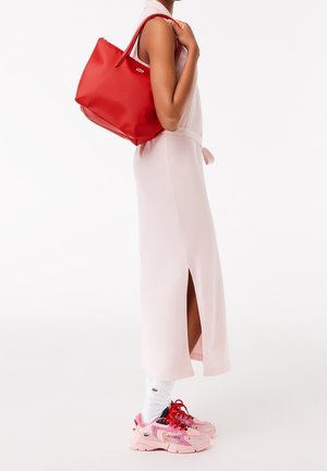 Vrouw in een lichtroze mouwloze jurk met zijsplit, witte sokken, roze en rode sneakers, met een grote rode draagtas over haar schouder.