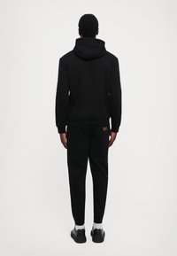 Man in een zwarte hoodie met bijpassende joggingbroek met het gouden EA7-logo, staand met zijn gezicht weg van een effen lichte achtergrond.