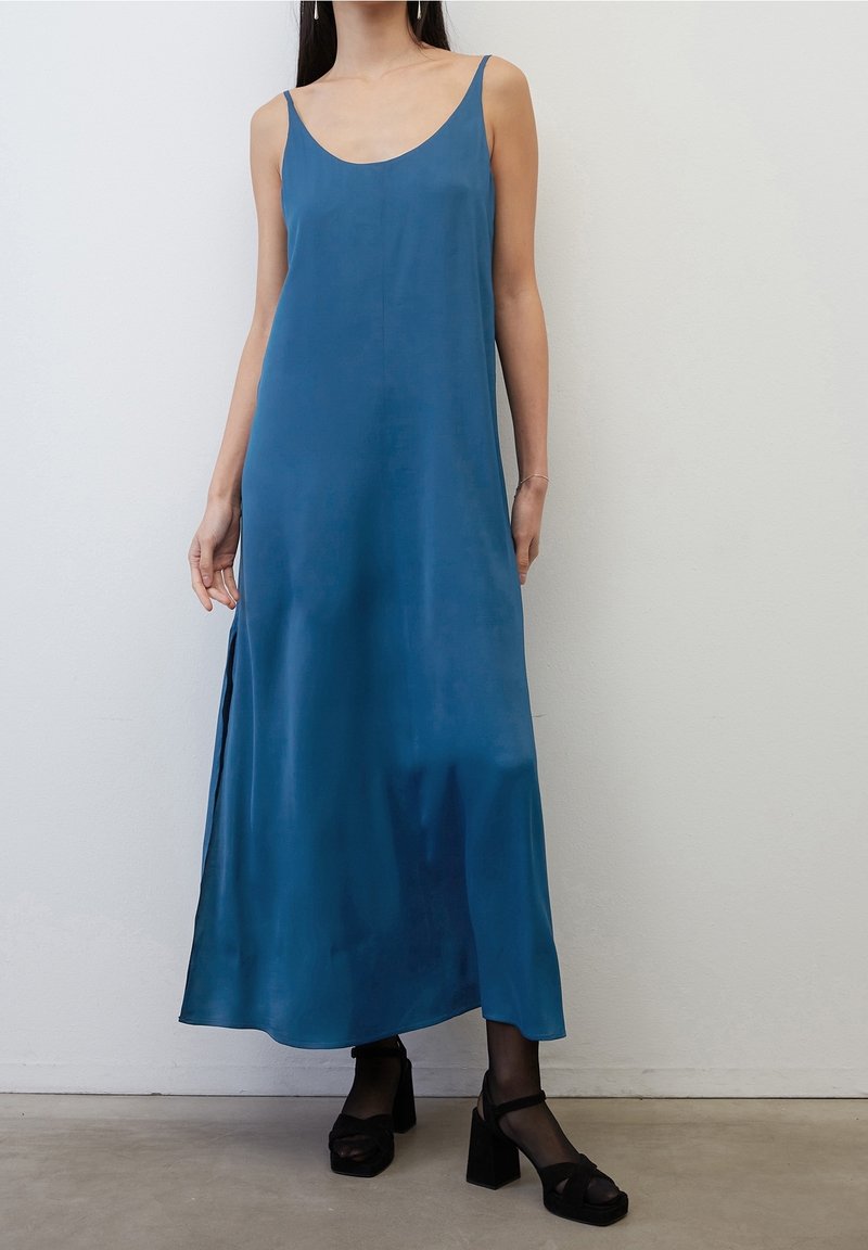 Femme portant une robe midi en satin bleu sans manches avec une fente latérale, des collants noirs transparents et des sandales noires à talons épais, debout contre un mur blanc.