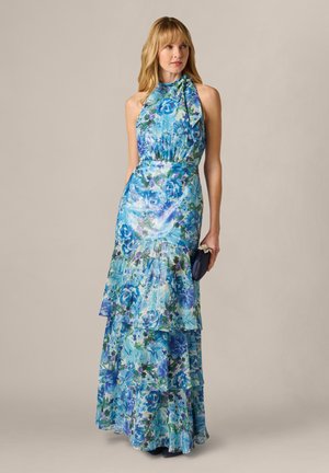 Kobieta w niebieskiej, kwiecistej sukience maxi z odkrytymi ramionami i wiązaniem na szyi, trzymająca małą ciemną kopertówkę, stojąca na jednolitym tle.