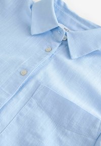 Chemise en lin bleu clair avec un col classique, deux boutons et une poche poitrine. Présente un tissu texturé et un motif à carreaux subtil.