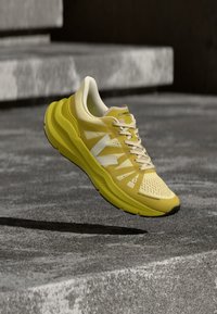 Κίτρινο αθλητικό παπούτσι με λευκές λεπτομέρειες και κορδόνια, αιωρούμενο πάνω από μια υφή τσιμέντου με σκάλες στο φόντο.