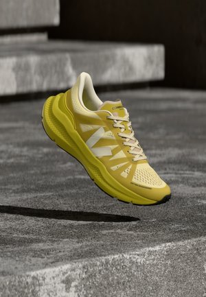 Pantof sport galben cu accente albe și șireturi, plutind deasupra unei suprafețe de beton texturată, cu trepte în fundal.