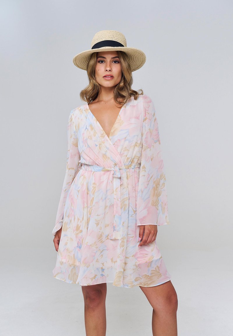 Femme portant une robe portefeuille fleurie pastel et un chapeau de paille, debout devant un fond uni gris clair.