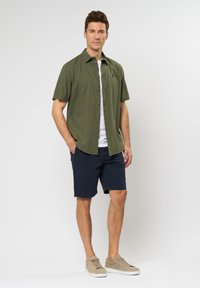 Camicetta verde oliva a maniche corte con abbottonatura frontale, abbinata a pantaloni corti blu navy e sneakers beige. Design semplice e materiale leggero.