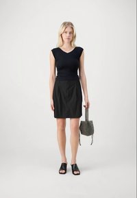 Blondinka stoji v brezrokavnem črnem topu, črni krilu, črnih odprtih sandalih z visoko peto ter drži majhno črno teksturirano torbico ob sebi.