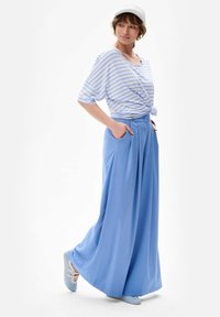 Gestreepte blauw-witte oversized shirt, geknoopt op de taille, gecombineerd met lichtblauwe, hoge brede broek en blauwe schoenen.