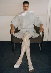 Grauer, übergroßer Sweatshirt mit hohem Kragen, schichtiger Rock, leggings mit Spitze und weißen Absätzen; sitzt auf einem Stuhl vor einem neutralen Hintergrund.