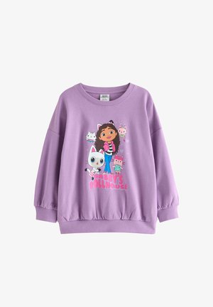 Enfant aux cheveux bouclés portant un pull violet et des leggings assortis avec des personnages de dessin animé et des baskets, debout sur un fond uni.