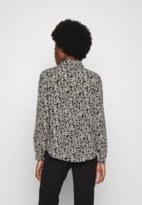 Camisa de manga longa em tecido preto com padrões florais brancos, apresentando um decote com colarinho e um corte descontraído, visível pela parte de trás.