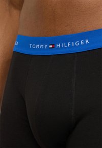 Μαύρα βαμβακερά αντρικά εσώρουχα τύπου μποξέρ με μπλε ελαστική ζώνη στη μέση που φέρει το όνομα της μάρκας "TOMMY HILFIGER" σε λευκό, συνοδευόμενο από έμβλημα λογοτύπου.