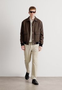 Casaco de lã castanho com fecho de botão e grandes bolsos, usado sobre uma camisa de tom claro e calças listradas em bege, com sapatos pretos.