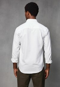 Weißes Langarmhemd mit hochgekrempelten Ärmeln, ausgestattet mit einem Button-Down-Kragen und einem geraden Saum. Getragen mit braunen Hosen, vor einem grauen Hintergrund präsentiert.