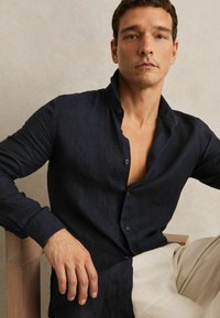 Hombre con camisa azul marino oscura desabrochada y pantalones beige, sentado en una silla de madera frente a un fondo liso.