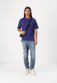 Camiseta de rayas púrpura con mangas cortas y un logo amarillo, combinada con jeans de un azul claro y zapatillas blancas, llevando un bolso cruzado negro.