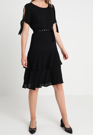 Vrouw draagt een zwarte jurk tot op de knie met een ceintuur in de taille, een geplooide rok en korte mouwen met strikjes, gecombineerd met zwarte pumps.