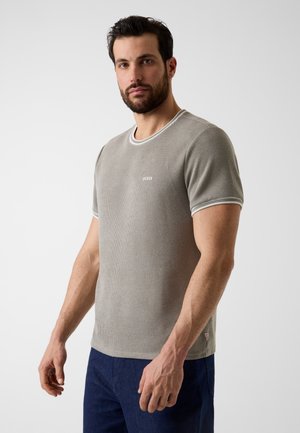 Man met baard, gekleed in een grijs gebreid shirt met korte mouwen en donkere broek, staand tegen een effen lichte achtergrond, iets schuin naar de zijkant gericht.