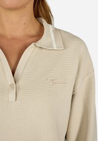 Šviesiai rudos vaflinio audinio polo marškiniai su kontrastinga apykakle, turinčia baltą juostelę, su mygtukų eilute ir siuvinėtu logotipu ant krūtinės.