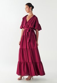 Dlouhé vínové šaty s překládaným designem, krátkými puff rukávy, vázaným pasem a vrstvenými volány na lemu. Hladká tkanina s jemnou texturou.