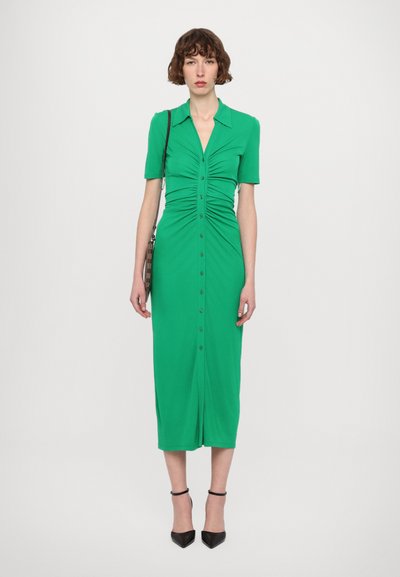 Vestido verde hasta la rodilla con mangas cortas, diseño de cuerpo fruncido, cierre frontal con botones y textura suave. Combinado con tacones negros puntiagudos.