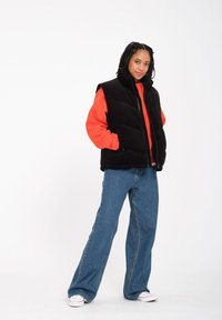Schwarze Puffer-Weste mit hohem Kragen über einem orangefarbenen Hoodie, kombiniert mit weit geschnittenen blauen Jeans und weißen Sneakers. Glatte und isolierte Textur.