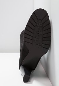 Bottine en daim noir avec une semelle en caoutchouc texturée, dotée d'un talon effilé et d'un motif de bande de roulement antid érapant pour une meilleure adhérence.